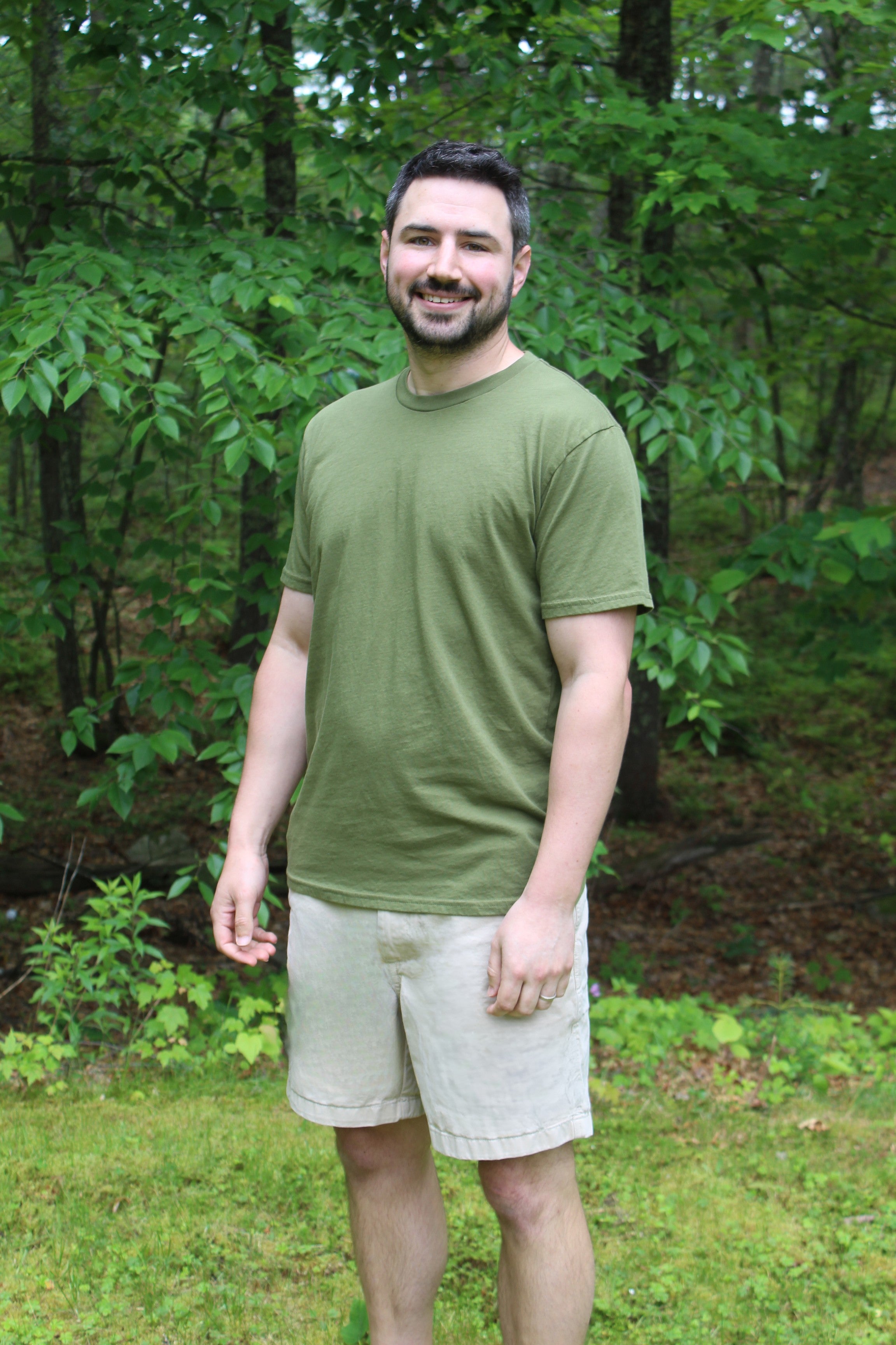 Man wearing a green organic cotton tee and shorts, standing in a green, outdoor setting surrounded by trees.